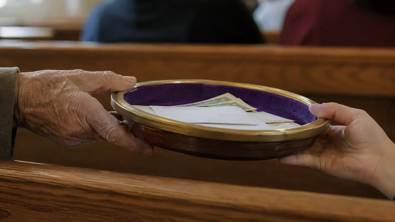 Two hands passing a wooden offering plate along a church pew