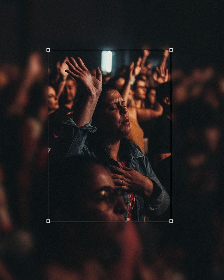 A woman in worship with one hand raised and one on her heart, framed by a thin white border, the congregation blurred around her